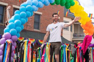 Bros — a man Man wearing white t-shirt and brown shorts stands under balloon rainbow waving into a crowd