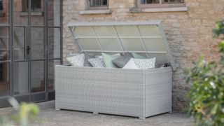 Light grey rattan-style outdoor storage box placed on a stone patio beside a rustic brick house. The box is open, filled with patterned and pastel cushions