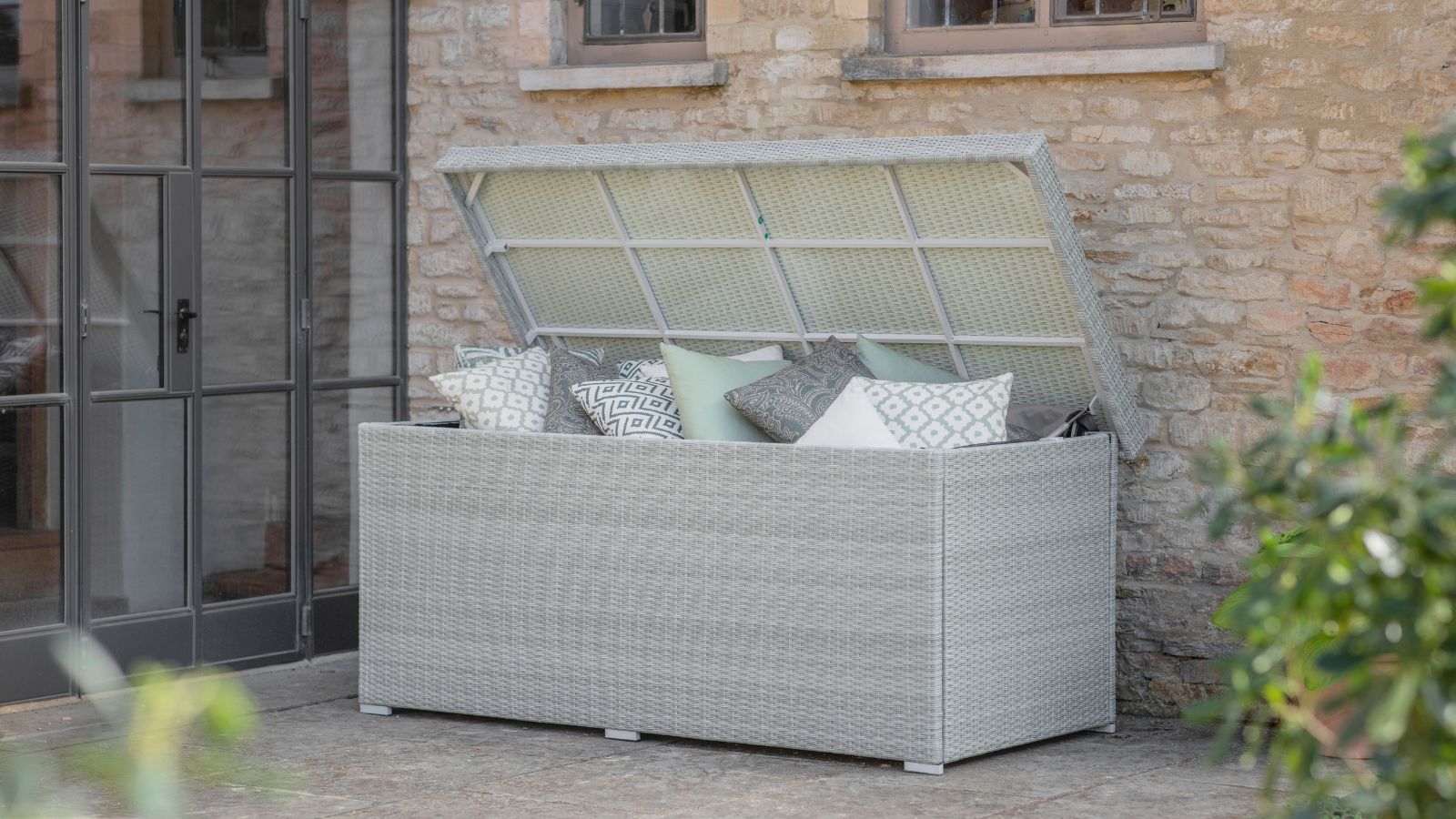 Light grey rattan-style outdoor storage box placed on a stone patio beside a rustic brick house. The box is open, filled with patterned and pastel cushions