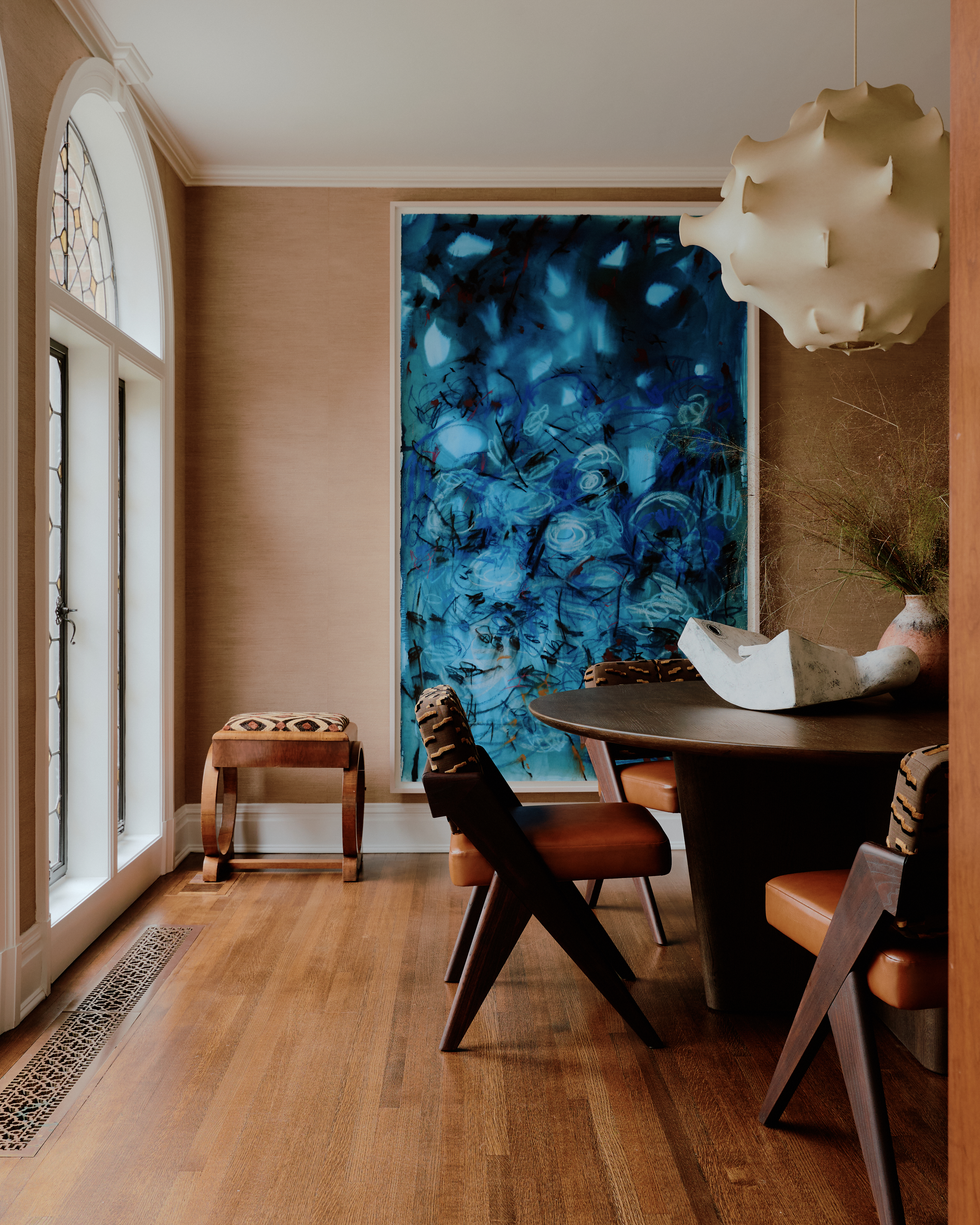Interior of a palatial residence with wooden floors, mid-century furniture, a sculptural pendant lamp in stretched fabric, and a large blue canvas hung up on terracotta walls.