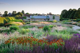 A flowers-filled field features bright patches of green, purple, red, pink, and orange, a series of bronze, stylized sculptures, and a house, all bathing in the sun.