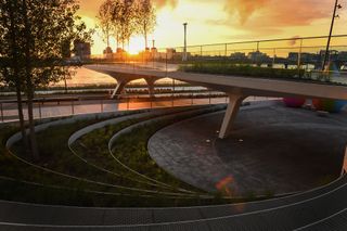 Alternative view of The Tide riverside park as the sun sets. The park features a raised walkway, greenery, trees and colourful waterdrop-shaped structures. The river can also be seen along with buildings on the other side
