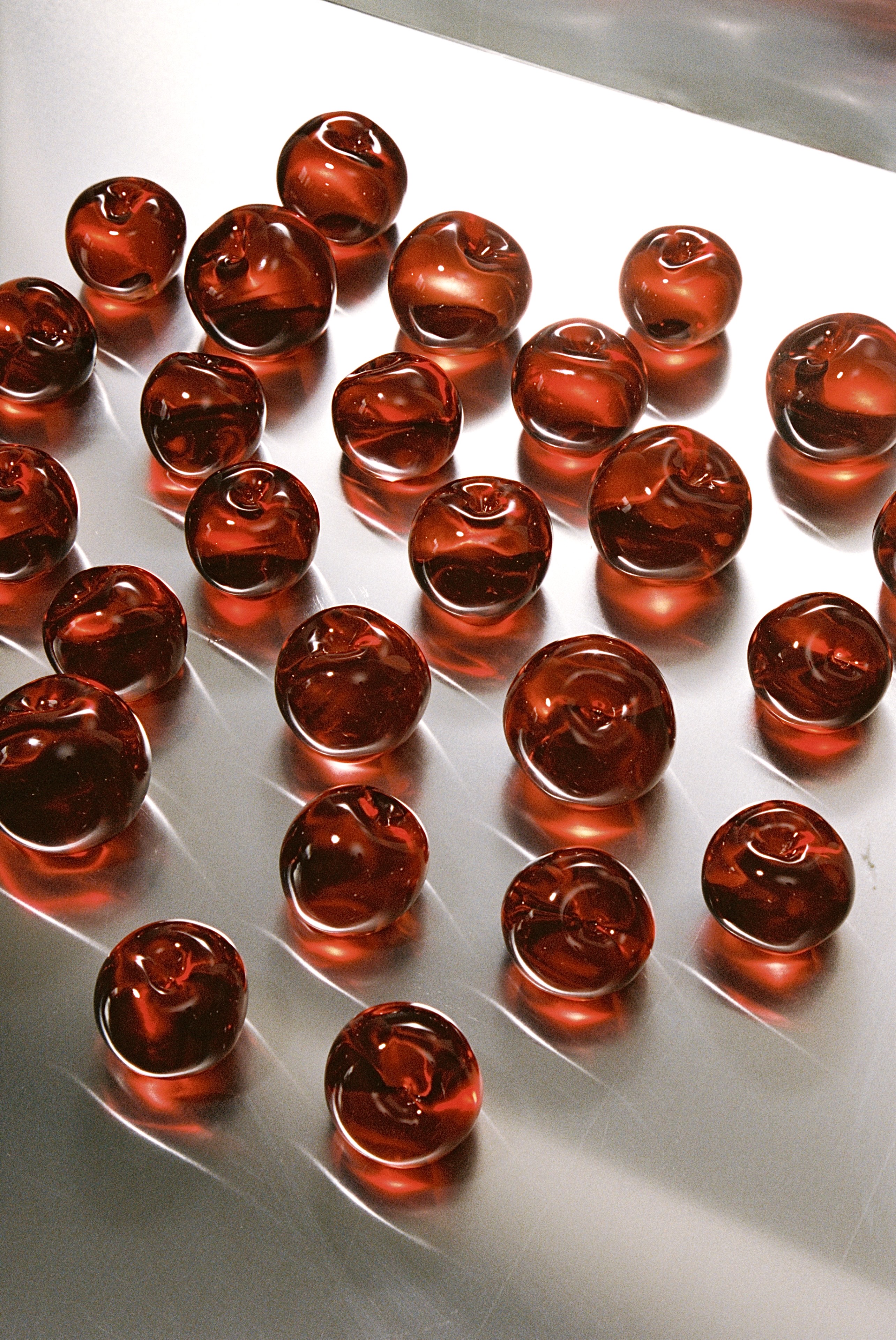 A series of glazed cherries stacked up on a chrome surface that reflects their gloss.