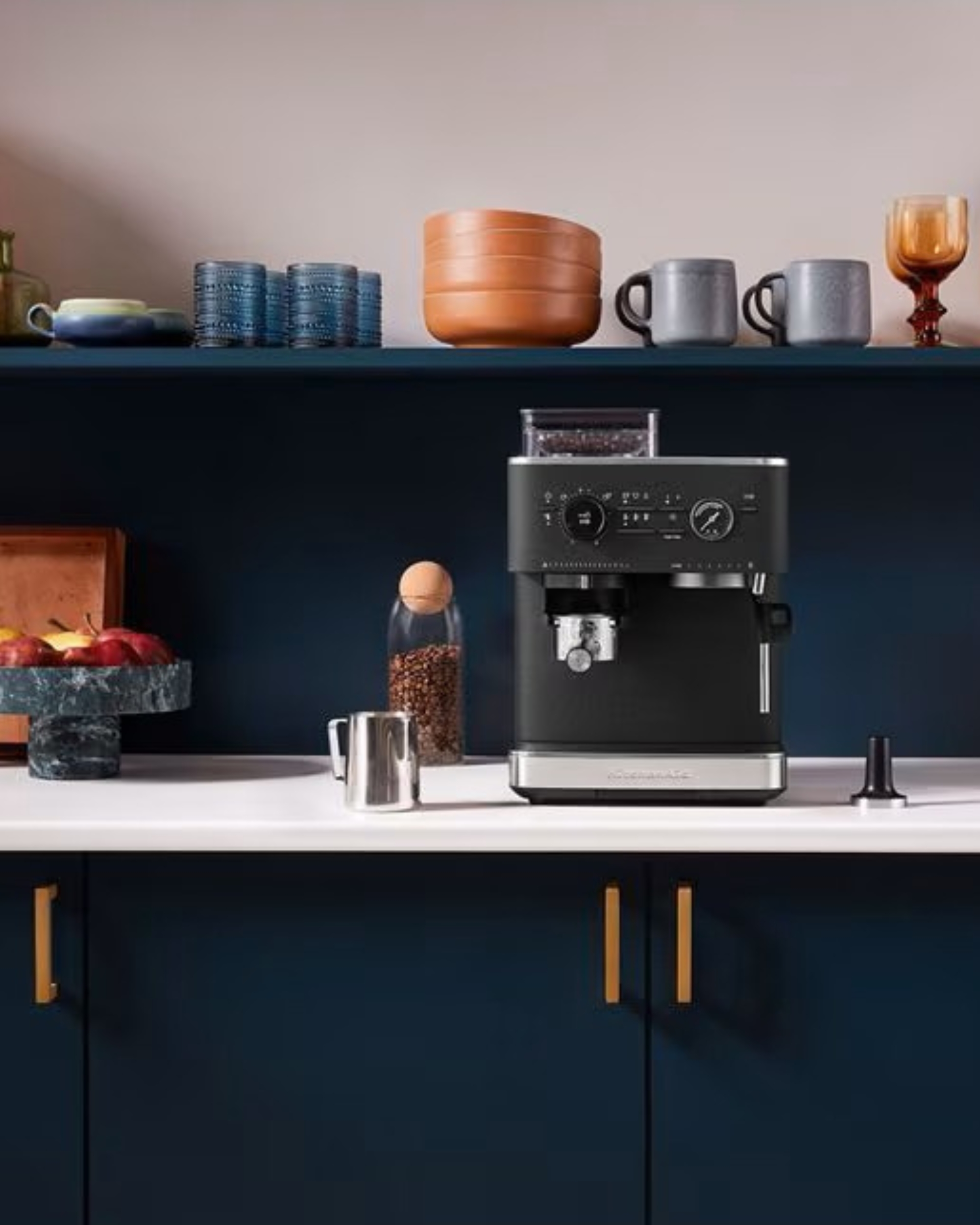 Image of a black espresso machine on a kitchen countertop. The countertop is whtie and the cabinets are a navy blue color with gold pulls. There is a a floating shelf with bowls and glasses stacked on it.