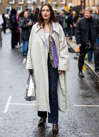 Alexa Chung wears a trench coat, a silver sequined bag, blue striped pants outside JW Anderson during London Fashion Week February 2024