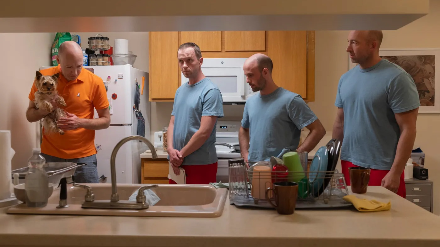 A man in an orange shirt holds a small dog, while surrounded by three men in blue shirts and red trousers in The Rehearsal, season 2, episode 3.