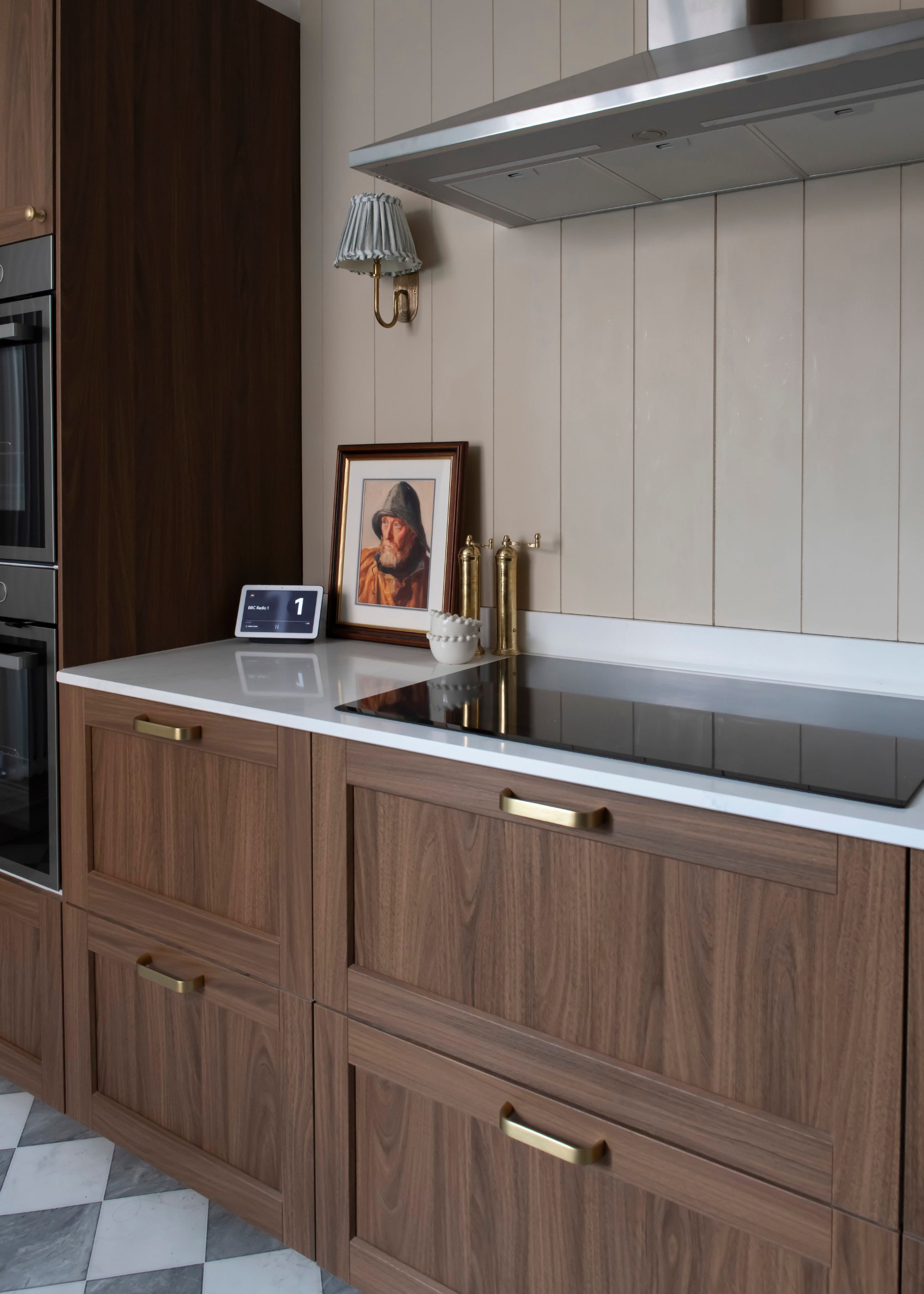 A dark wood kitchen with drawers and gold hardware on the drawers. There is also an induction stovetop with an extractor fan above it. On the white quartz kitchen countertop is a salt and pepper mill, a painting and a smart calender/clock.