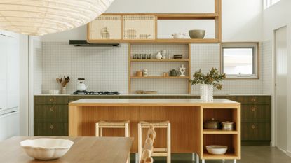 A green and white kitchen with an open plan, a wood island, overhead rattan-style shelving, a white tile backsplash, a paper pendant lantern and a wood dining table