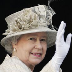 Queen Elizabeth wearing a beige hat with flowers and matching coat with white gloves and waving against a black background