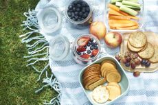 A picnic blanket is on the grass with fruit vegetables and crackers on it. 
