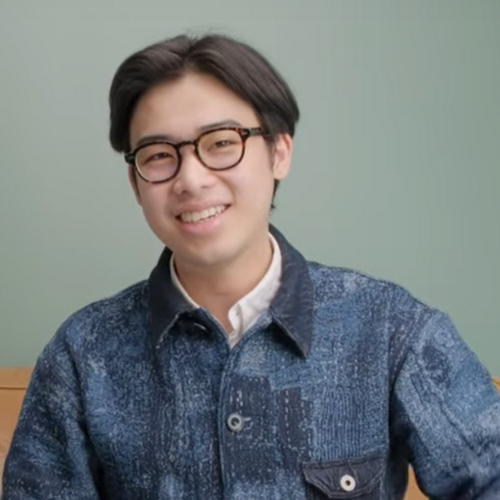 Image of a man in glasses with a navy blue collared shirt-jacket. He is sitting in front of a sea moss green background.