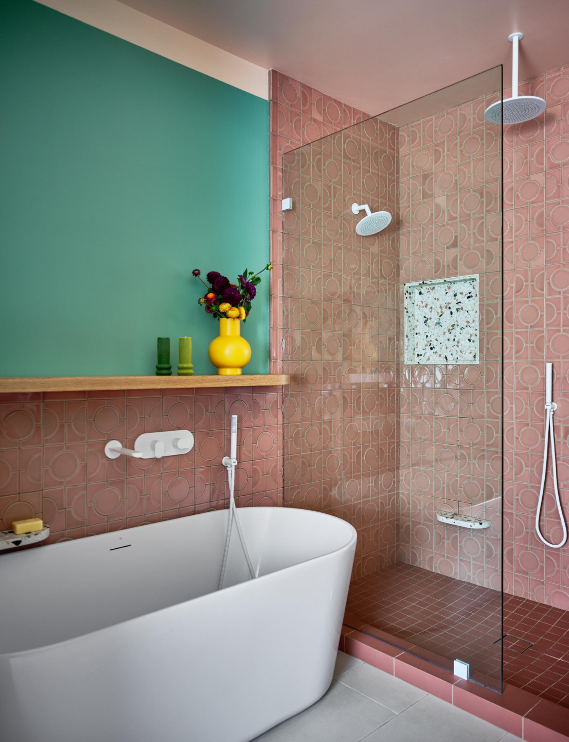 Glass shower enclosure with geometric pink wall tiles, a deep rose mosaic floor, white fixtures and a tiled curb beside a freestanding bathtub.