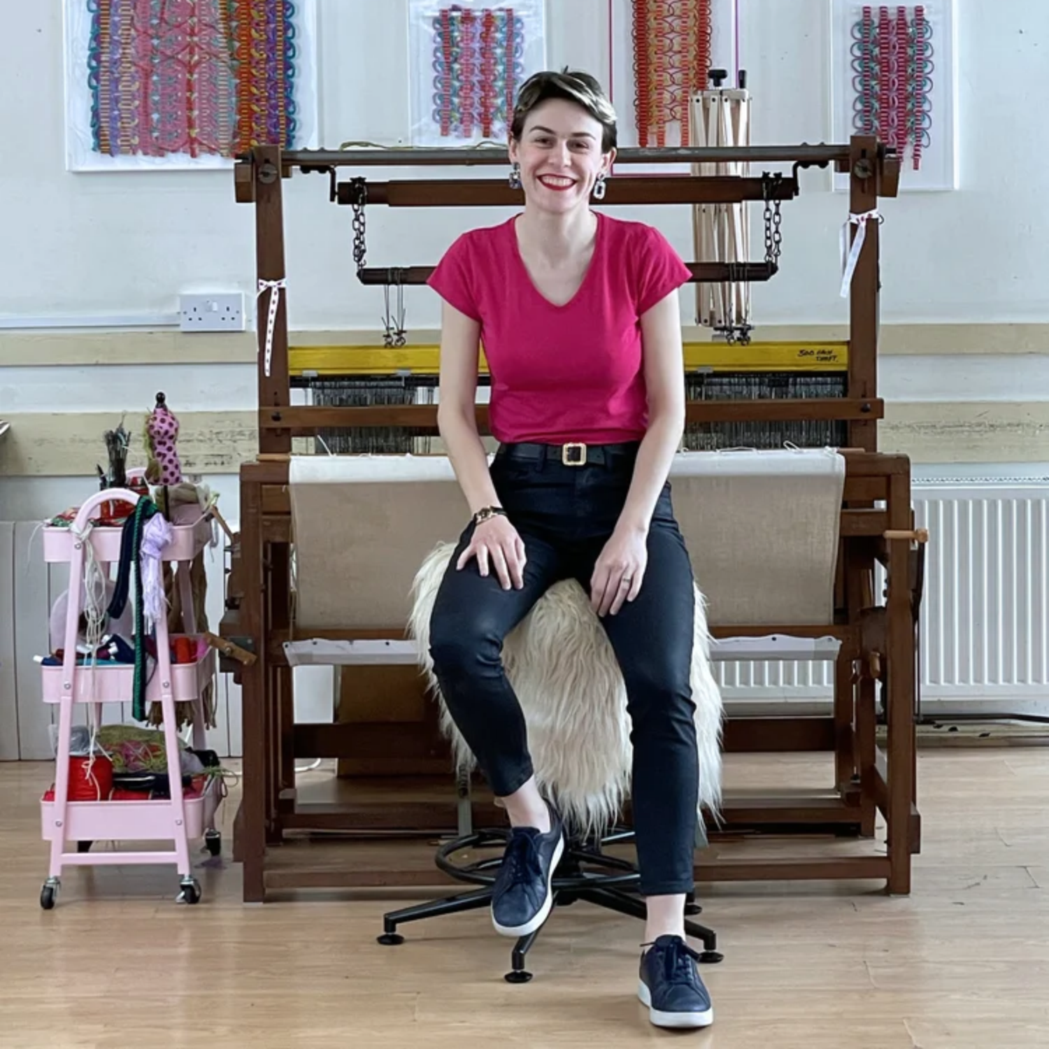 A woman on a fuzzy chair in a passementerie studio, wearing a pink top and black jeans