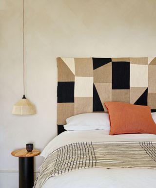 Bedroom with textured, cream painted walls, headboard with natural, black and cream woven design, white bed linen with orange cushion and patterned throw, black and wood bedside table, low hanging pendant light with cream fabric shade.