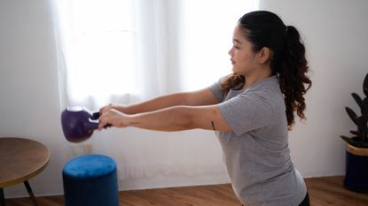 Woman performing a kettlebell swing in a domestic setting. She wears a grey T-shirt