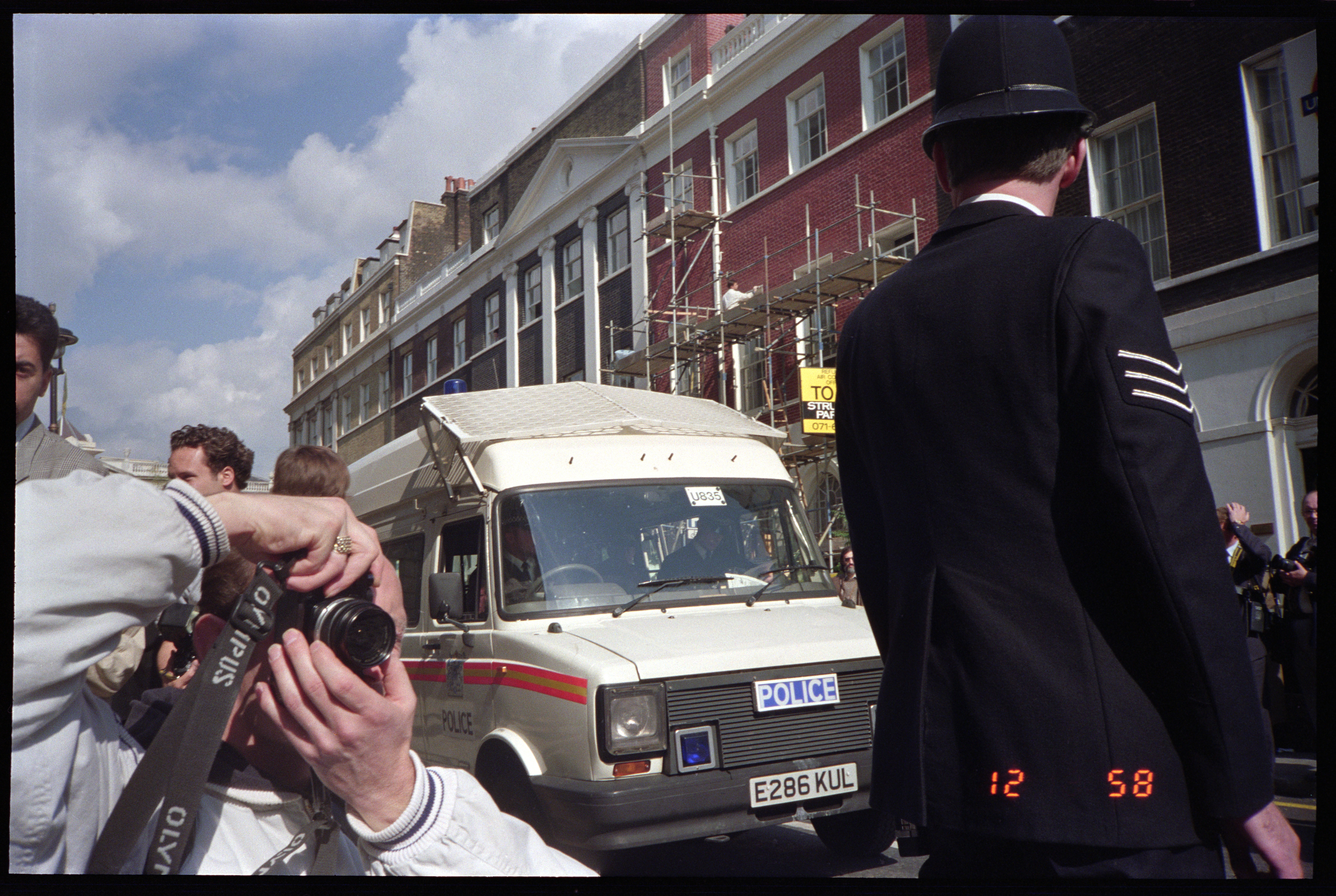 A photographer crouches low to shoot a Metropolitan Police van with an Olympus camera on a London street, a uniformed officer standing in the foreground.