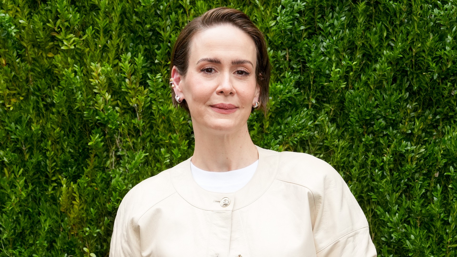 a white woman with short hair wearing a cream sweater and posing in front of a grass wall