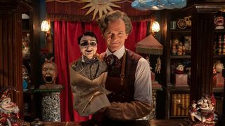 Neil Patrick Harris as The Toymaker, holding a creepy ventriloquist's dummy in a dimly-lit study full of spooky curios, in Doctor Who The Giggle, episode 3 of the 60th Anniversary trilogy.