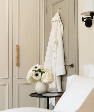 A white bathrobe hanging on the back of a sage green painted door in a panelled bedroom. A black bed side table with a small round vase of white flowers.