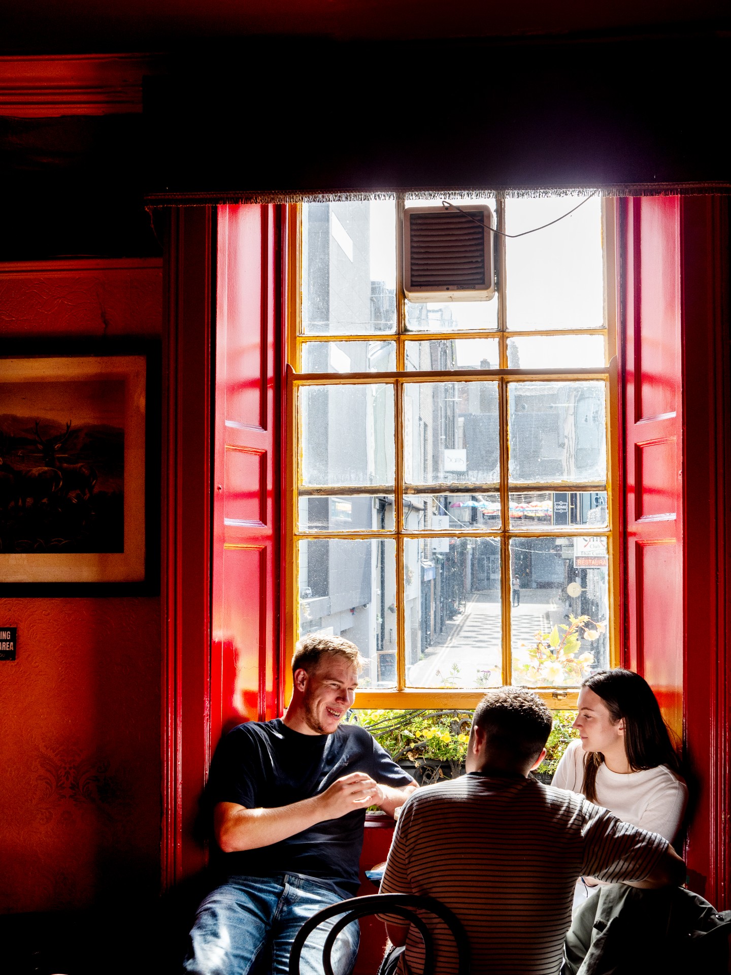 Three young friends chat at a table in front of a bright sash window framed by glossy red shutters in a dimly lit, red-walled pub, with a narrow street visible outside.