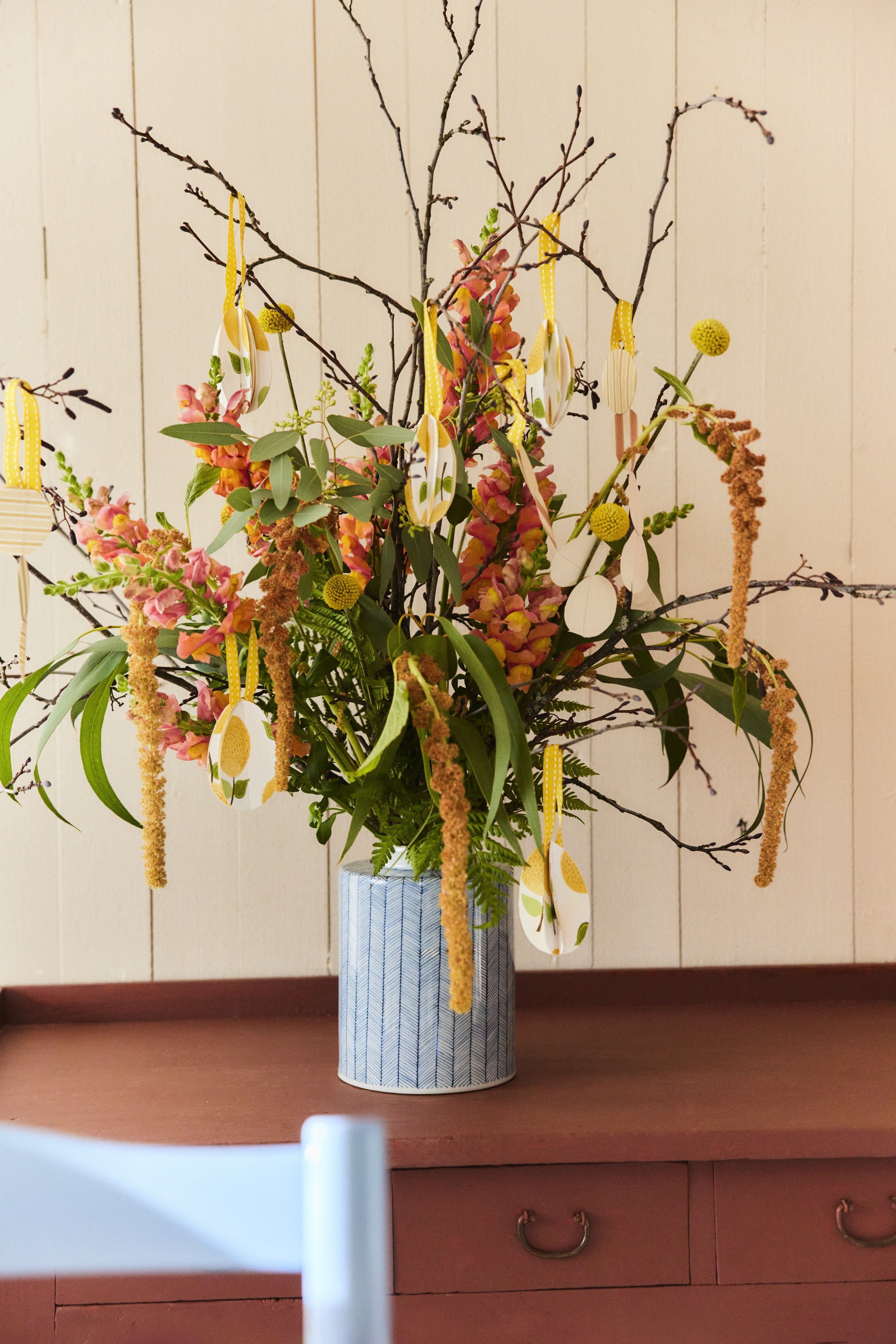 Image of a pink and orange floral arrangement in a light blue vase sitting on a brick red cabinet with white walls behind it. There are ornaments made out of wallpaper hanging from the stems.