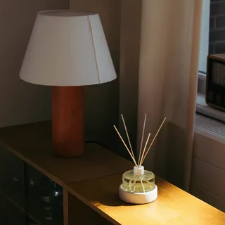 A reed diffuser sits atop a warm slab of wood decorated with an organic table lamp.