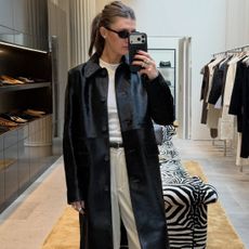 NYC style influencer Coco Schiffer posing for a mirror selfie wearing black oval sunglasses, a black collared leather coat, white T-shirt, a black belt, and cream trousers