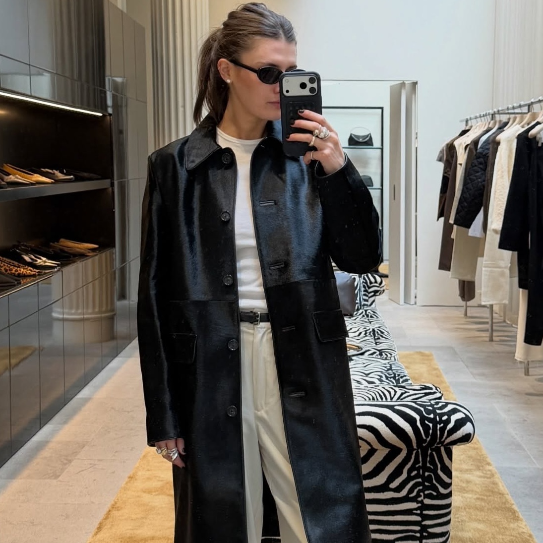 NYC style influencer Coco Schiffer posing for a mirror selfie wearing black oval sunglasses, a black collared leather coat, white T-shirt, a black belt, and cream trousers