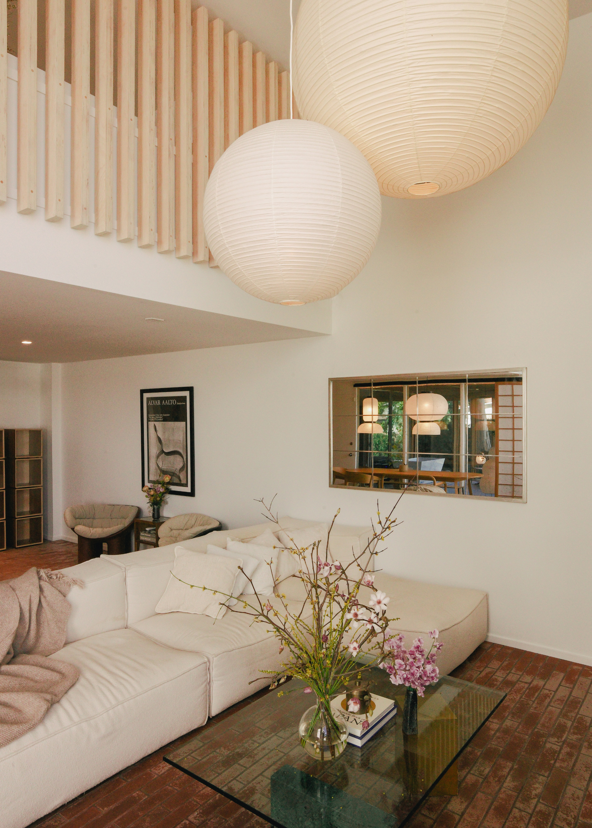 A living room with a cream sectional sofa, a glass coffee table with vases of flowers, and a pair of rice paper lamps hanging from above
