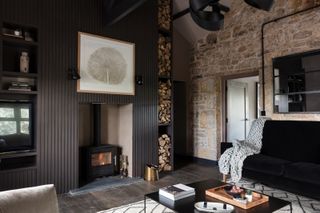 Living room with original brick wall, large fireplace with woodburning stove surrounded by dark painted slatted panelling. Black velvet sofa and sleek black square coffee table