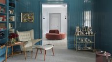 Image of a living room drenched in a blue lacquer paint color. There is a white mid-century chair and table by the built-in bookshelf on the left side of the room, and a gold bar cart on the right side of the room.