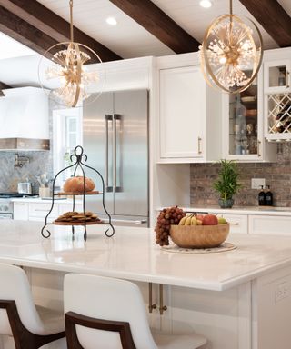 A white kitchen island with a tiered stand with bread and cookies on it and a wooden fruit bowl, two white chairs underneath it, plus white cabinets with gold handles and a silver two door fridge in front of it