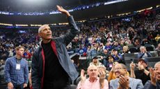 Barack Obama, in a black tee shirt and a blue coat, waves to fans at a basketball game