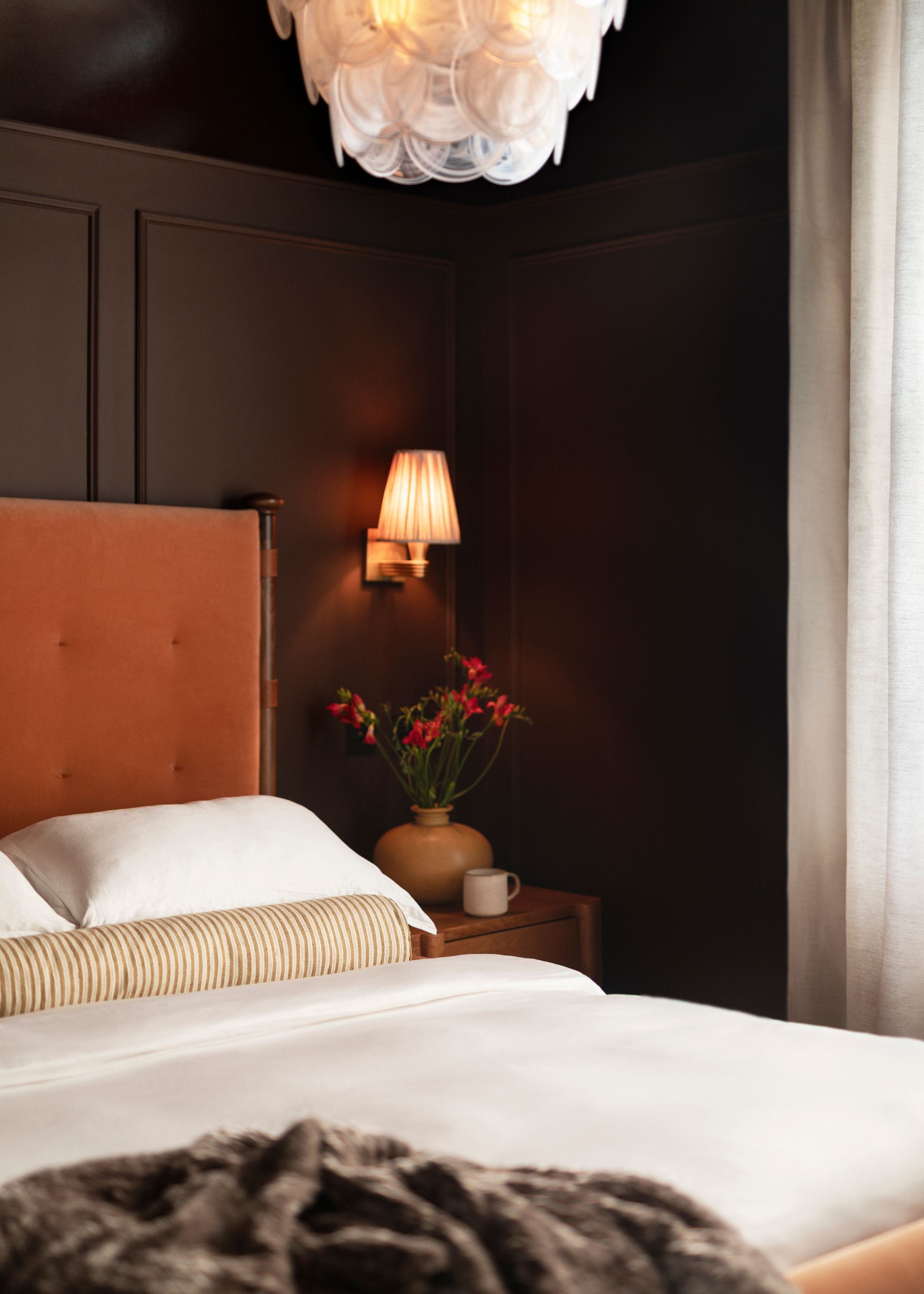 A brown bedroom with a Murano-style chandelier, warm wall sconce lamps, a bed with a tall headboard, white bedding, a striped bolster pillow, and a fuzzy throw beside a nightstand with a vase of flowers and a mug