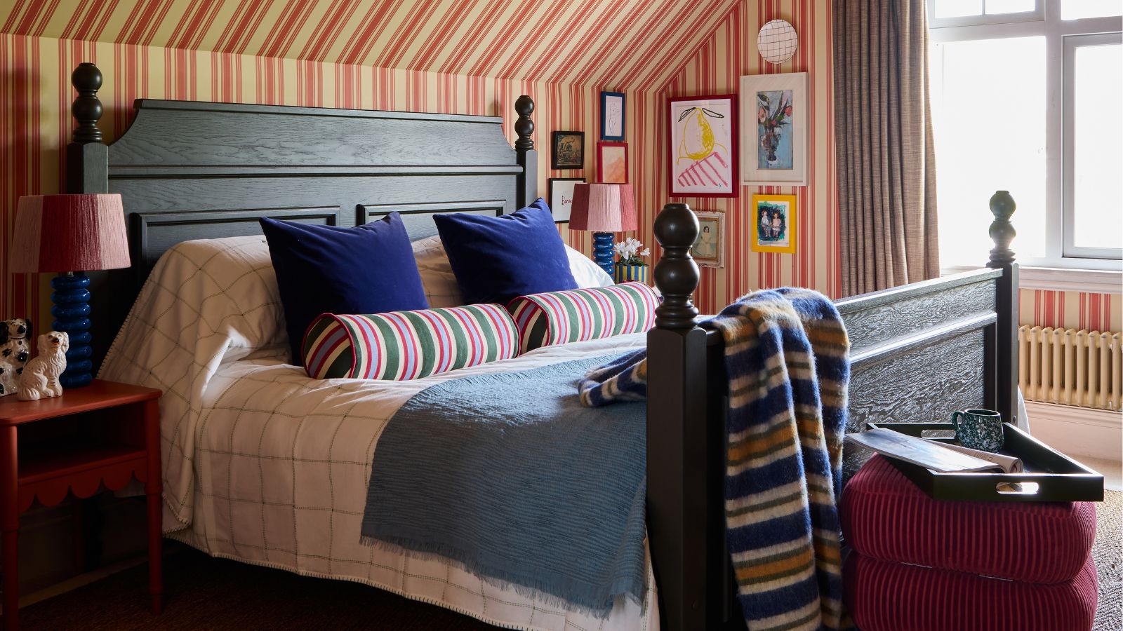 bedroom with a sloped ceiling and beige and red striped wallpaper that goes up and over the ceiling. A large dark wooden vintage bed sits in the middle styled with striped throws and pillows with a velvet stool at the foot of the bed