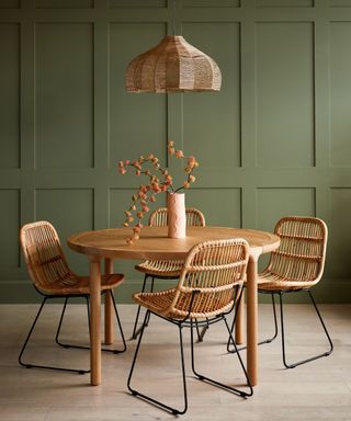 A boho dining room with dark green paneled walls, a circular wooden dining table with four rattan chairs around it, a vase of yellow flowers, and a curved bamboo pendant light above it and light wooden flooring below