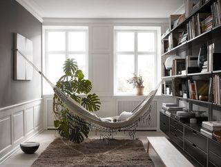 View of the library area with a hammock at The Apartment gallery. The space features wood flooring, grey walls with white wood panelling at the bottom, two windows, radiators, a green plant, a rug and a black wooden unit that has shelving filled with books and drawers at the bottom