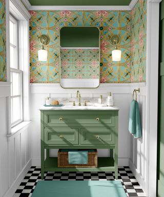 A green small bathroom with gold and blue patterned wallpaper, two gold wall sconces and a curved gold mirror, a standing green vanity with a marble countertop, and black and white checked flooring
