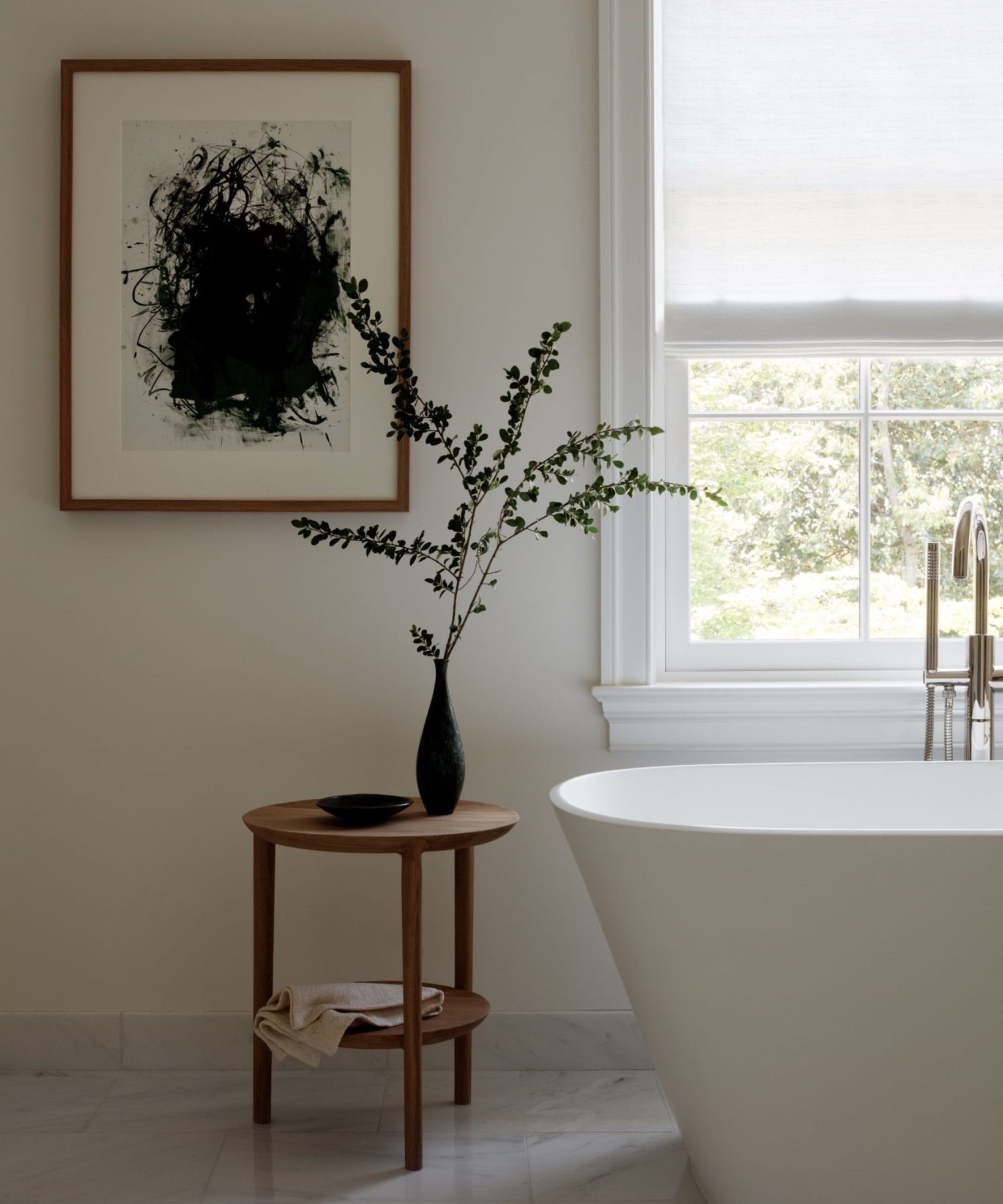 A minimalist bathroom with a modern freestanding tub, a dark wood side table, and abstract art on the wall