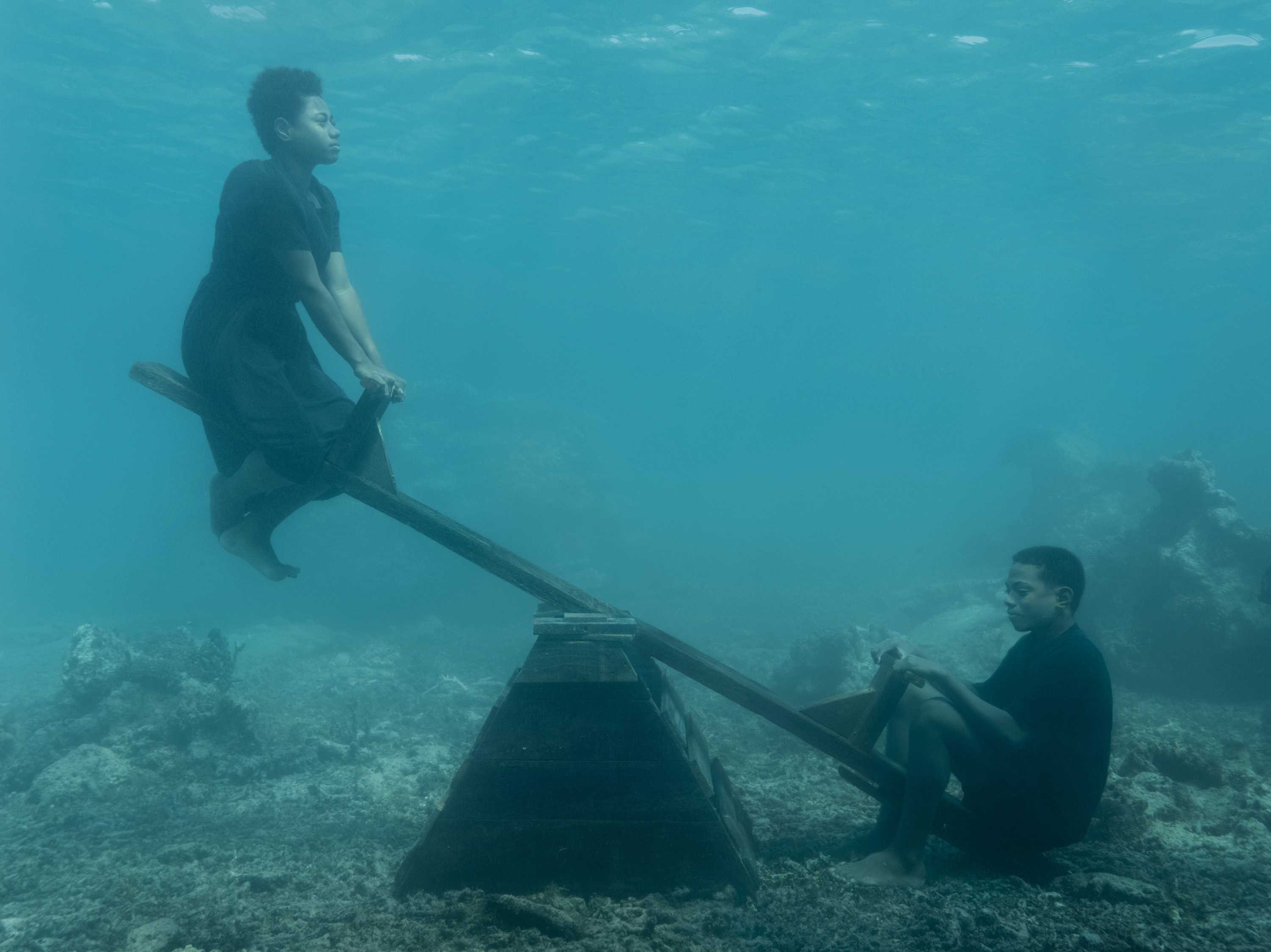 Two people sit on a wooden seesaw resting on the seabed, captured in a serene underwater environment.