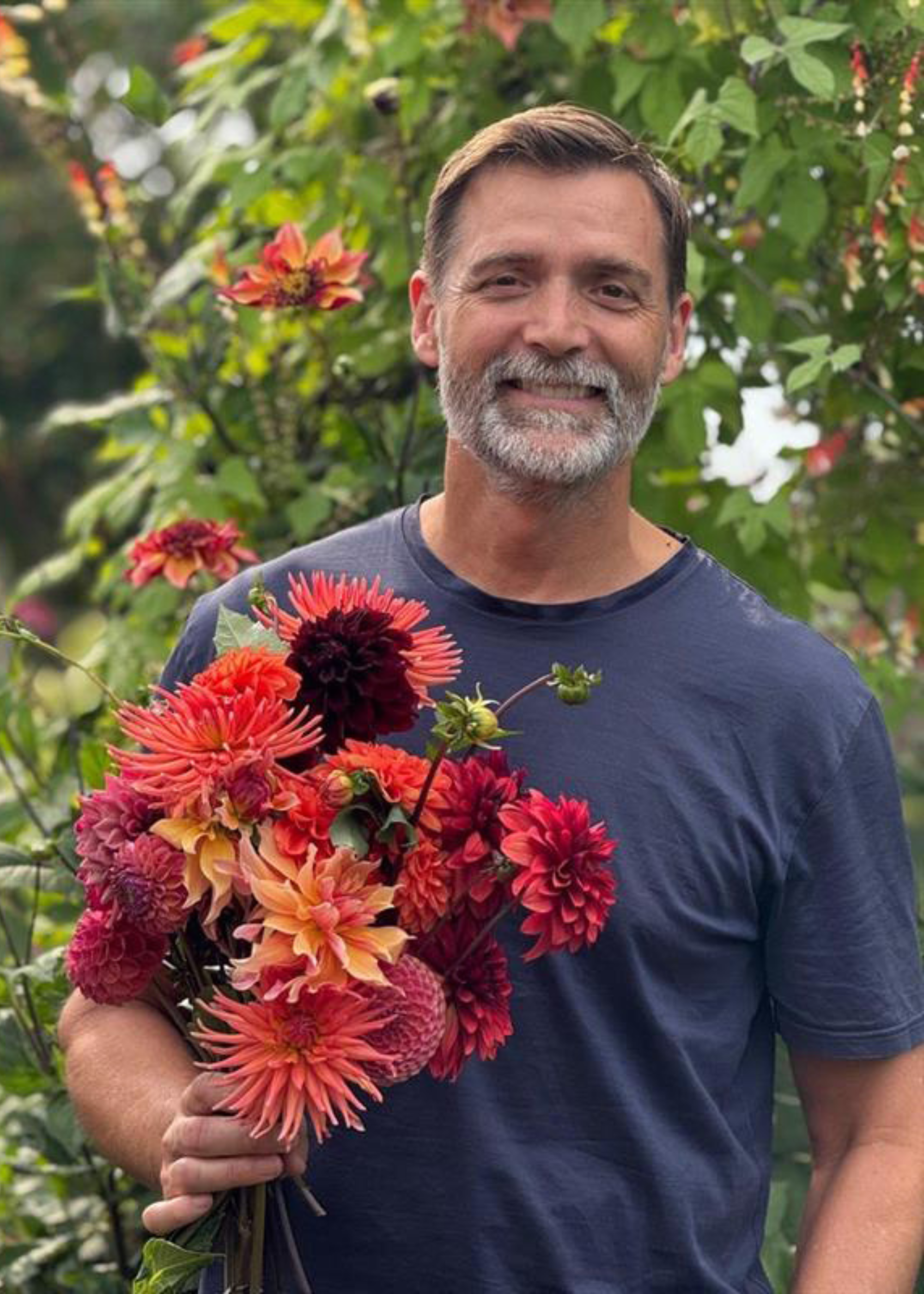 A man in a blue round-neck t-shirt holding colorful orange, pink, and burgundy dahlias