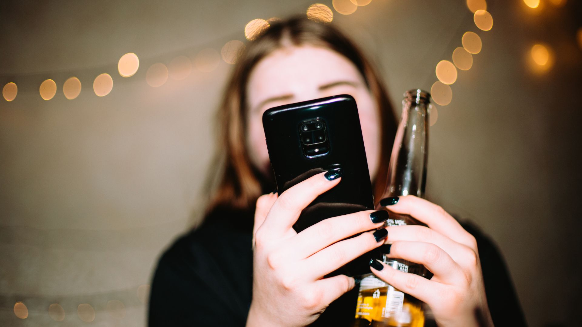 A woman looks at her phone, which is in front of her face, as she holds a beer.