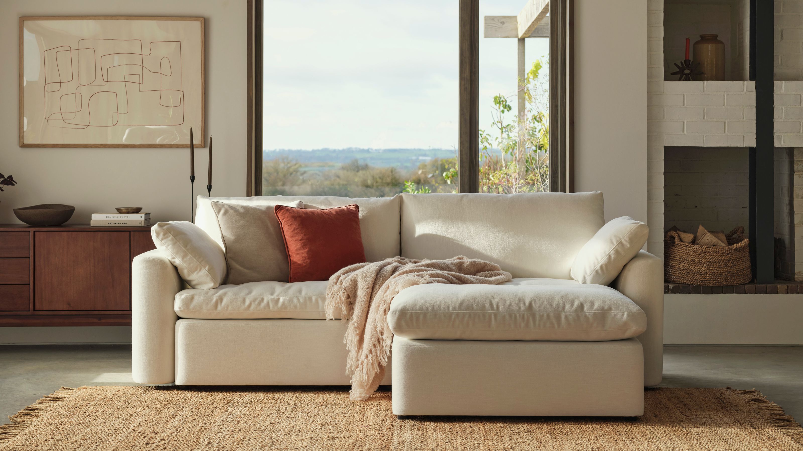 Swyft cloud sofa in a modern home with polished concrete floors, a jute rug, white walls with art and a timber console, a window looking out to a landscape, a built-in fireplace with shelves holding logs