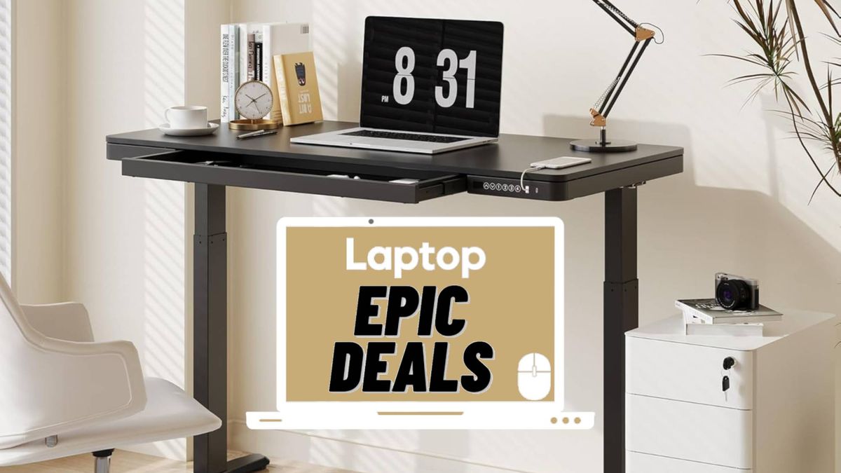 Silver laptop on a black Flexispot Comhar standing desk with phone charging via its USB slot surrounded by a desk clock, coffee cup and saucer, books, a desk lampt