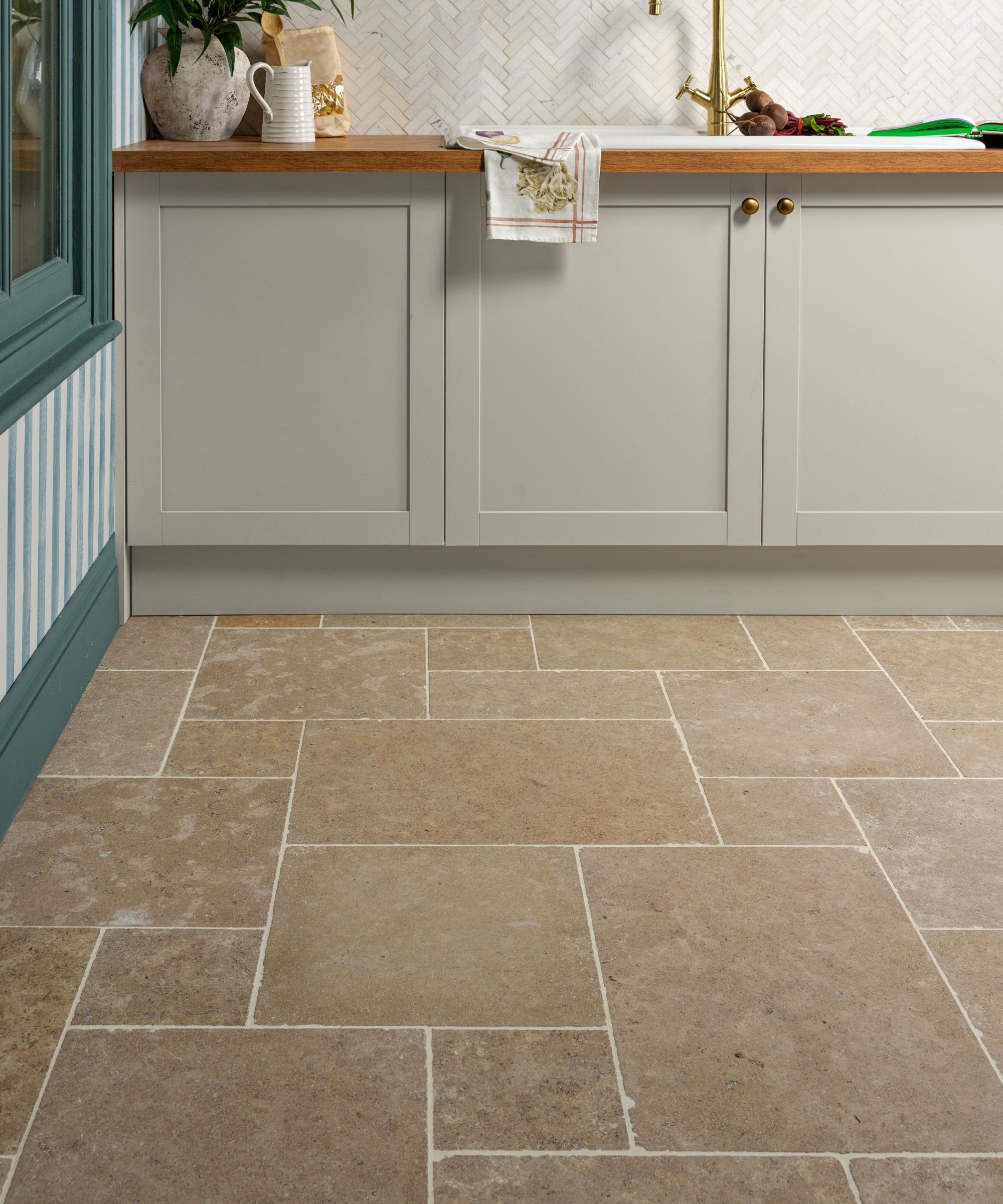 Cream shaker-style cabinets with brass knobs sit above warm, rustic stone floor tiles laid in a staggered pattern.