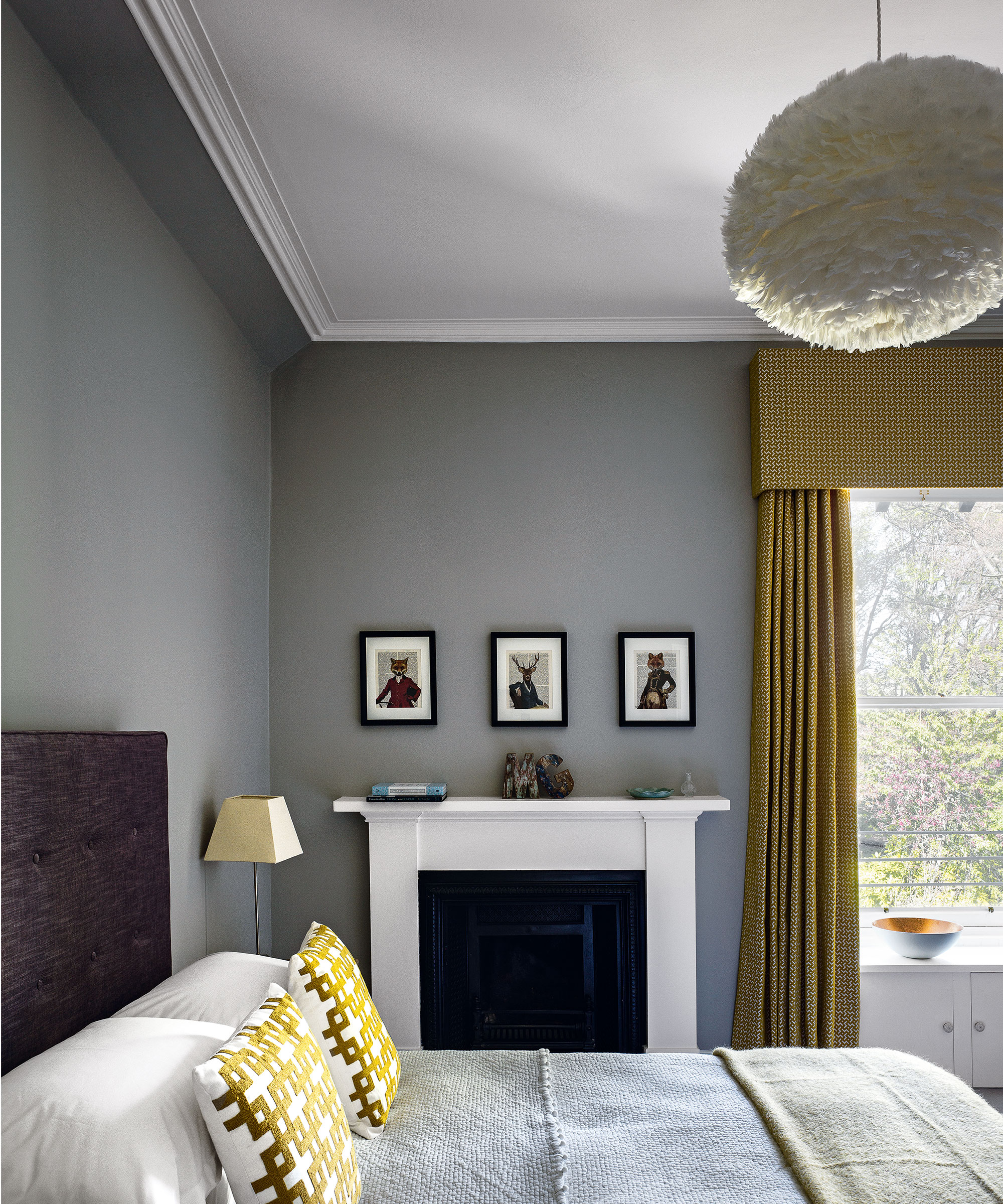 A bed with a dark grey headboard with gold decorative cushions in front of a traditional white fireplace and a bedside lamp
