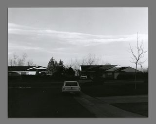 A black and white photograph of a residential street in Colorado showing an old white car on a driveway