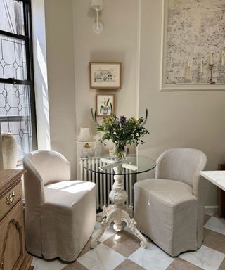 A dining area with two gray seats, a glass table, beige walls with wall art on it, and a white and dark gray checkerboard floor