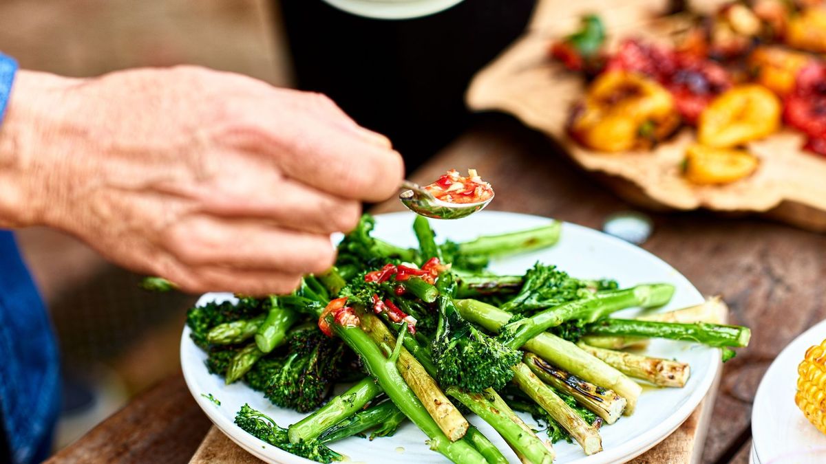 close up of an older white man&#039;s hand as he uses a spoon to garnish a plate of broccolini with a sauce. Other vegetable dishes can be seen in the background of the image.