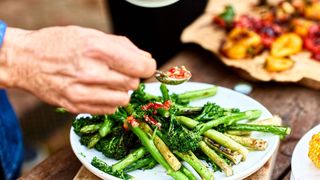 close up of an older white man's hand as he uses a spoon to garnish a plate of broccolini with a sauce. Other vegetable dishes can be seen in the background of the image.